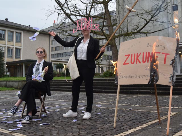 Die JUSO Baselland bei ihrer Aktion vor dem Finanzdepartement BL. Inszeniert wird eine reiche Person (links), die von Anton Lauber (mitte) mit Geld beworfen wird. Dieser hält zugleich eine Wurst über ein brennendes Schild, auf dem «Zukunft» steht. Foto: zVg